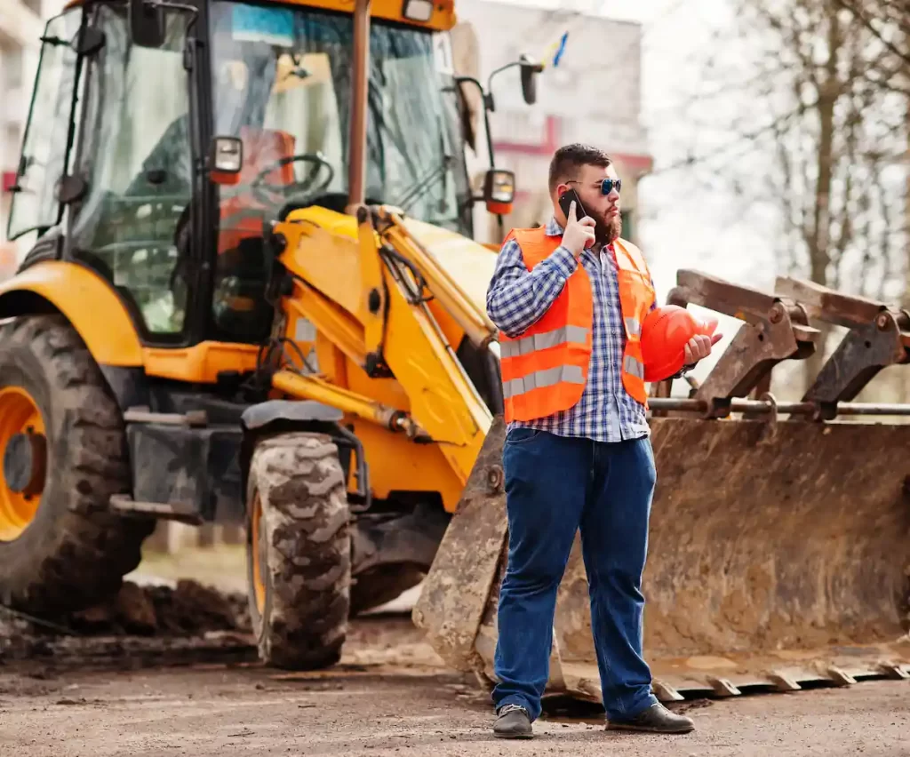 beard worker man suit construction worker safety orange helmet sunglasses against tractor with mobile phone hand